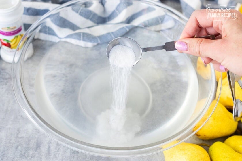 Pouring fresh fruit preserver from a measuring spoon into a large bowl of water. Pears on the side.