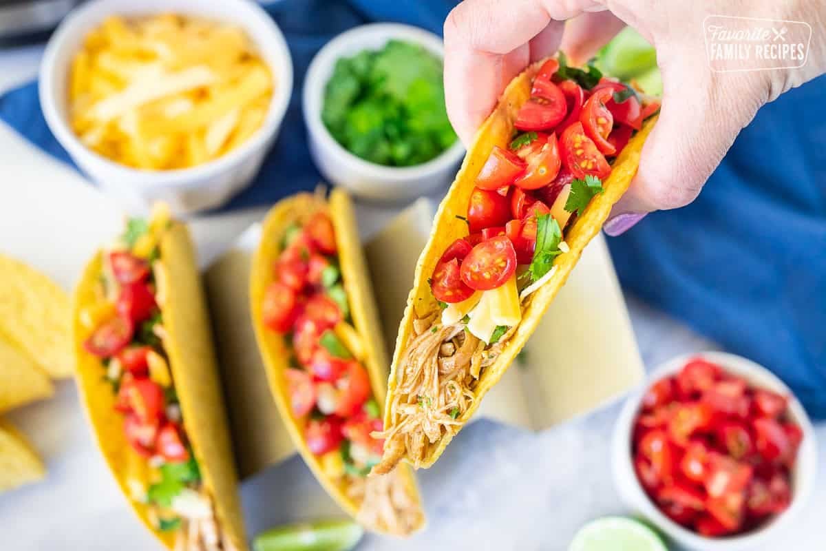 Hand holding a crunchy instant pot chicken taco topped with cilantro, cheese and diced tomatoes.