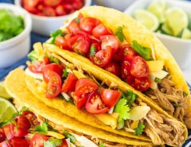 Three instant pot chicken tacos topped with cilantro, cheese and diced tomatoes on a plate. Limes wedges, tomatoes an cheese on the side.