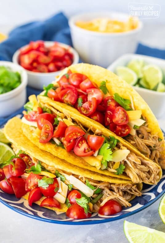 Three instant pot chicken tacos topped with cilantro, cheese and diced tomatoes on a plate. Limes wedges, tomatoes an cheese on the side.