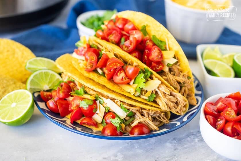 Three instant pot chicken tacos topped with cilantro, cheese and diced tomatoes on a plate. Limes wedges, tomatoes an cheese on the side.