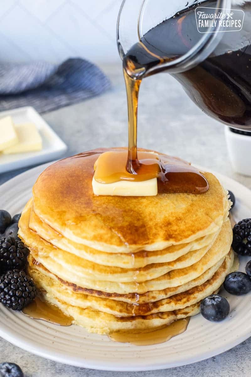 Pouring syrup onto a stack of pancakes from scratch on a plate with a pad of butter. Blackberries and blueberries on the side.