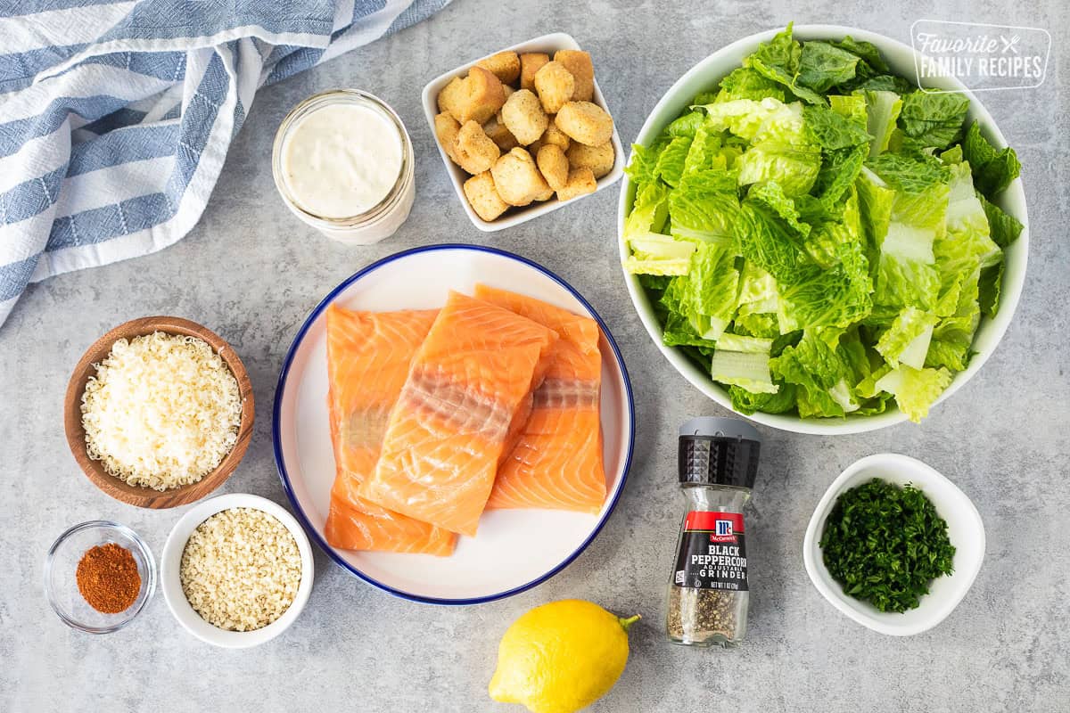 Ingredients to make Salmon Caesar Salad including chopped romaine lettuce, croutons, parsley, black pepper, lemon, salmon, caesar salad dressing, cheese, panko and paprika.