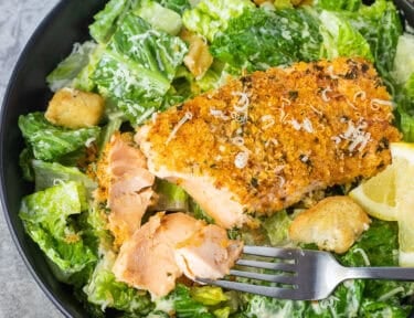 Broken up Salmon Caesar Salad with romaine lettuce, croutons, lemon wedges and fork.