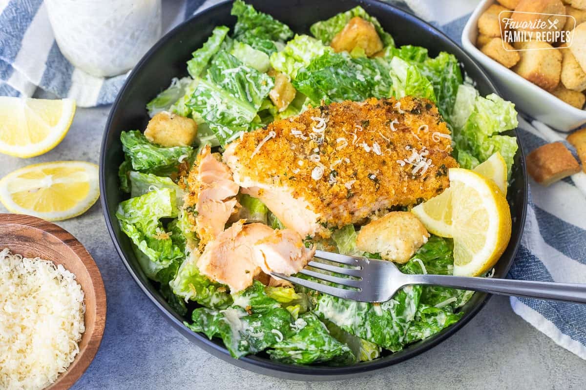 Broken up Salmon Caesar Salad with romaine lettuce, croutons, lemon wedges and fork.