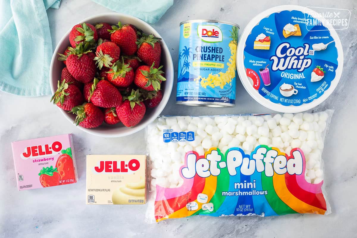 Ingredients to make strawberry fluff salad including Cool Whip, mini marshmallows, crushed pineapple can, strawberry Jell-O mix, vanilla pudding mix, and fresh strawberries.