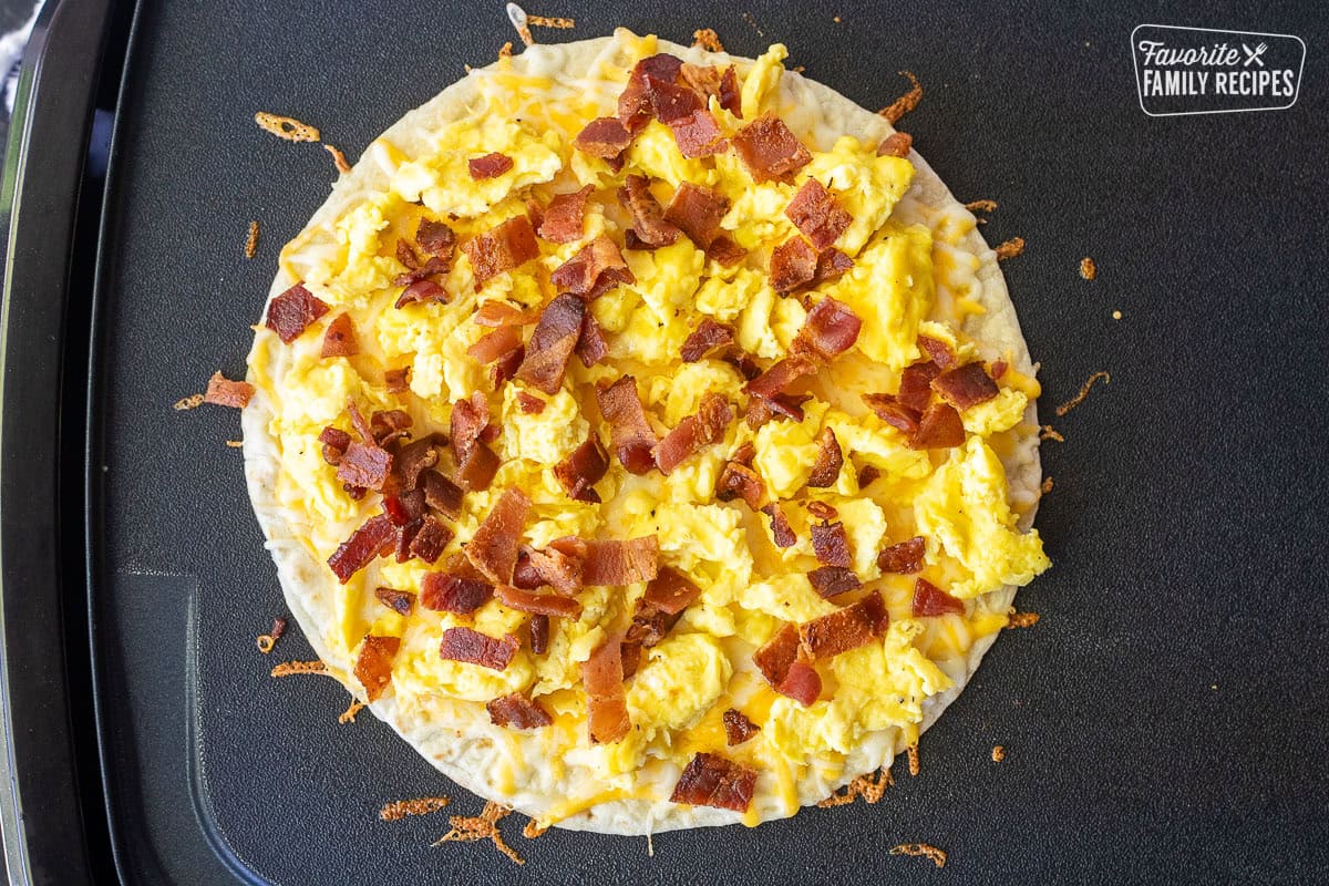 Griddle with tortilla, melted cheese, scrambled eggs and bacon pieces on top.