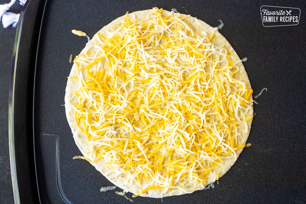 Griddle with flour tortilla and shredded Mexican blend cheese on top.
