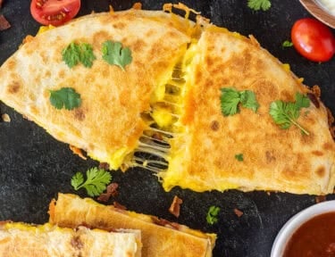 Breakfast Quesadilla pulling pieces apart with the melted cheese stretching. Garnished with cilantro and tomatoes. Sour cream and salsa on the side.
