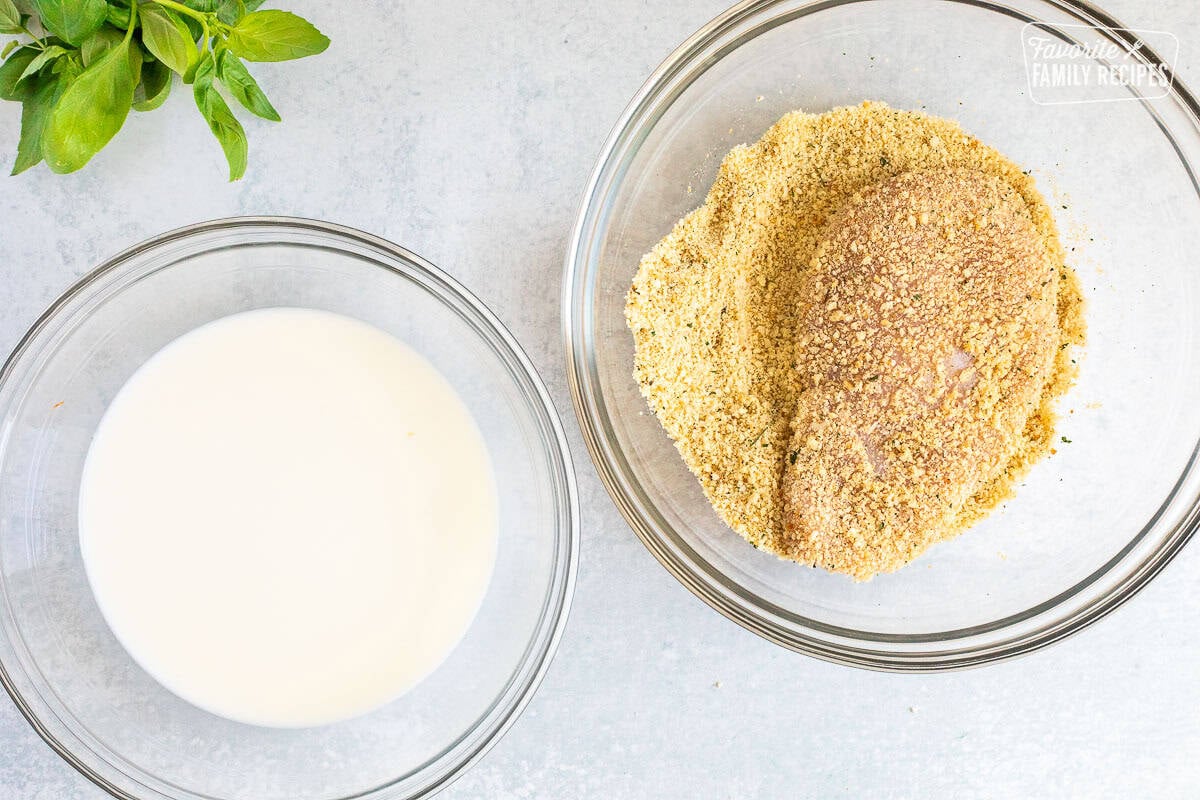 Chicken breast in a bowl of breadcrumbs next to a bowl of milk.