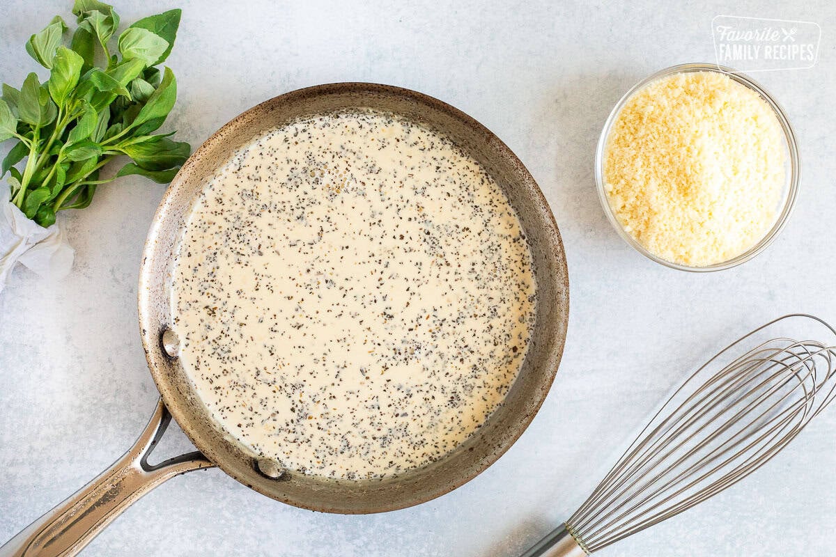 Skillet with creamy basil sauce next to a small bowl of Parmesan cheese, fresh basil and a whisk.