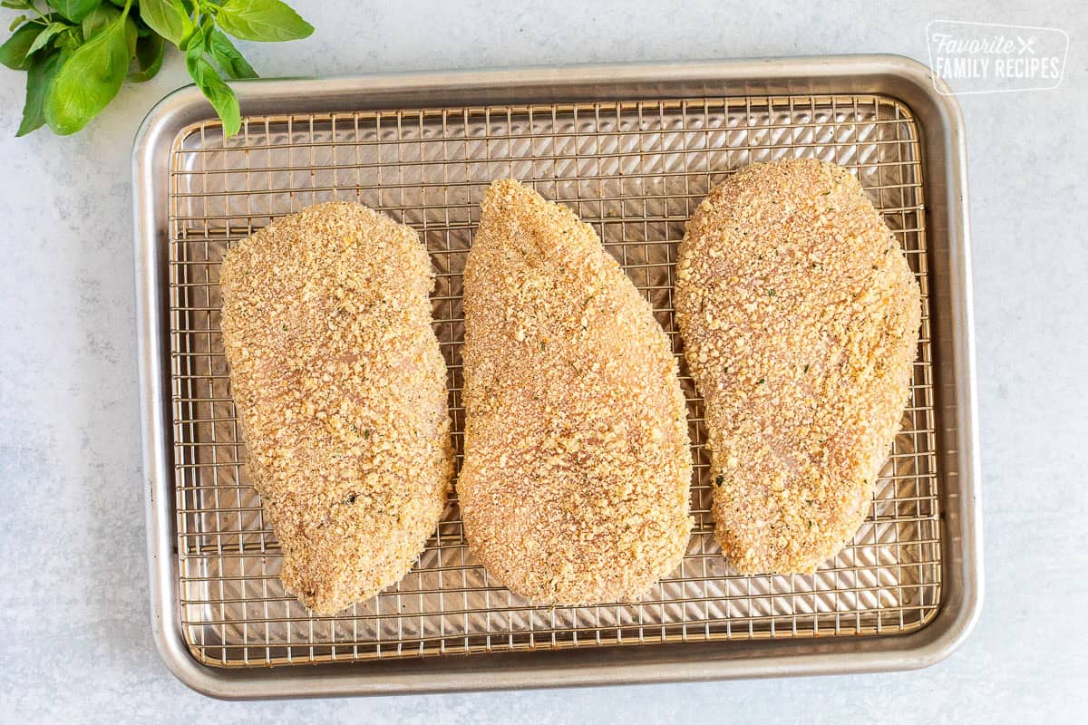 Three chicken breast breaded on a sheet pan rack.
