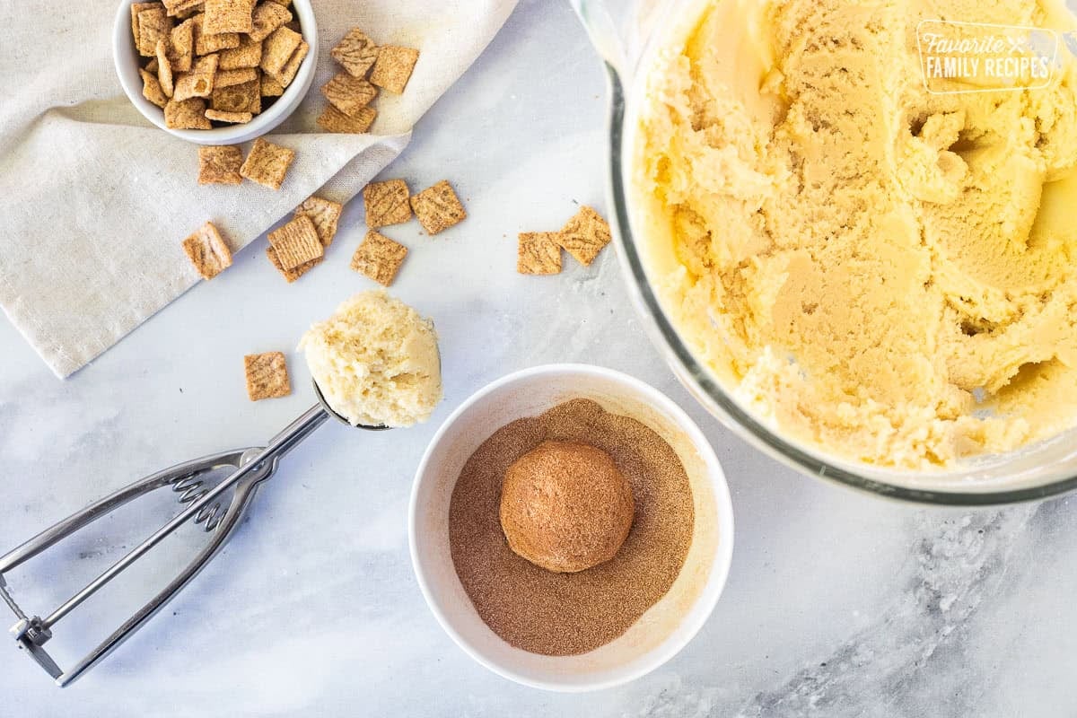 Glass mixing bowl with cookie dough, bowl of cinnamon sugar with a ball of covered cookie dough, cookie scoop with cookie dough.