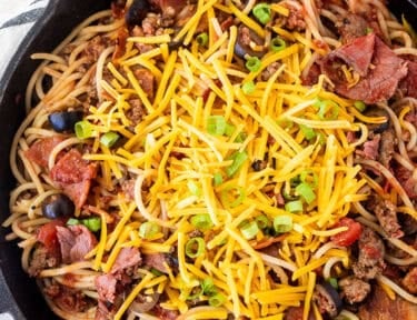 Skillet of cowboy spaghetti with noodles, ground beef, pastrami, bacon, pepperoni, olives, mushrooms, tomato sauce, cheddar cheese and green onion garnished on top.