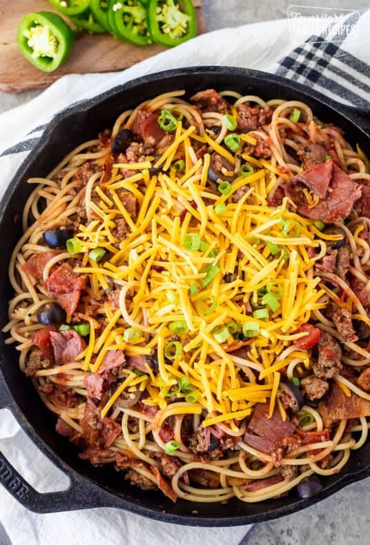 Skillet of cowboy spaghetti with noodles, ground beef, pastrami, bacon, pepperoni, olives, mushrooms, tomato sauce, cheddar cheese and green onion garnished on top.