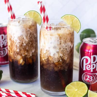 Two glasses of Dirty Dr Pepper with lime juice, coconut syrup and half and half on top. Straws and lime wedges on the sides of the glasses.