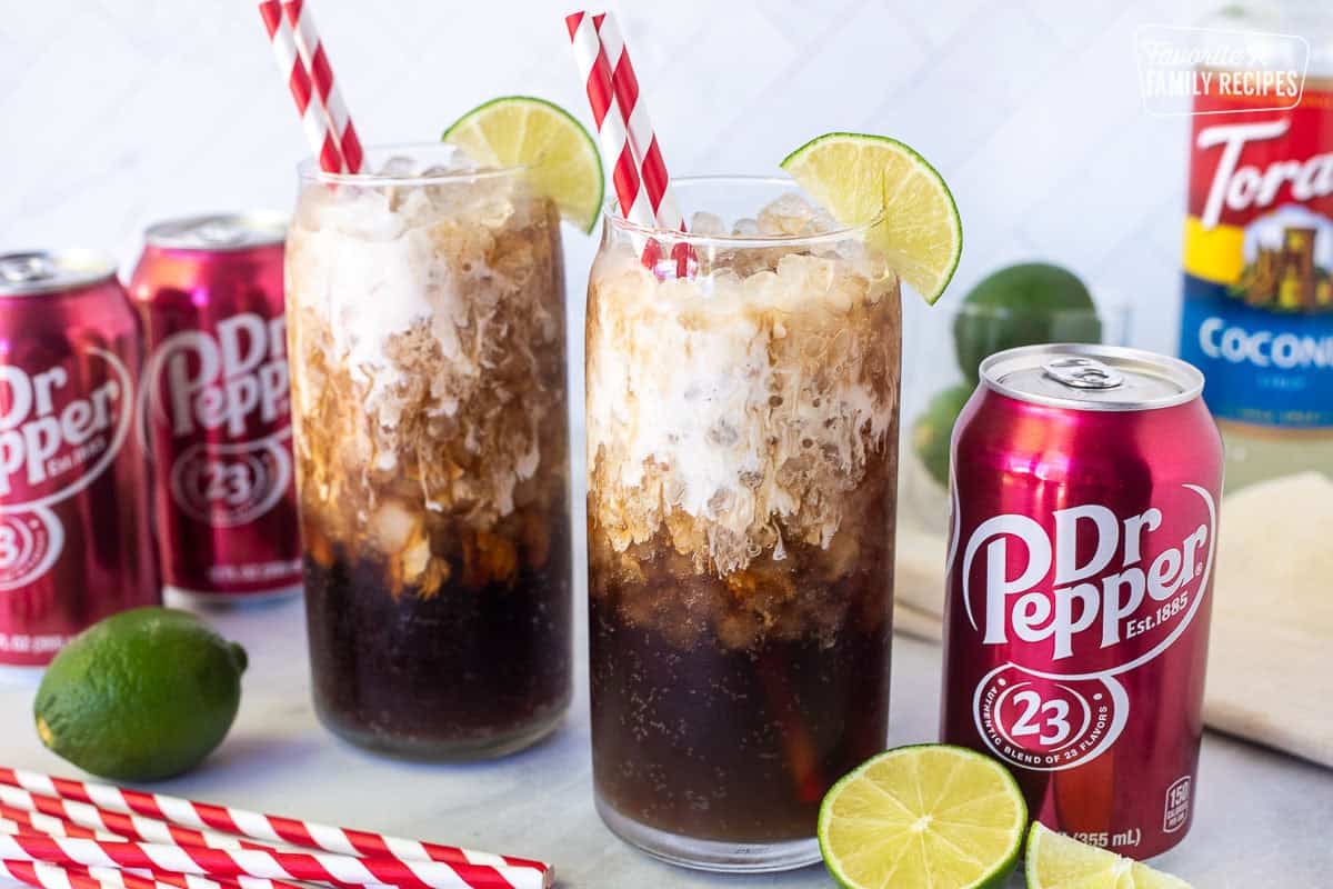 Two glasses of Dirty Dr Pepper with lime juice, coconut syrup and half and half on top. Straws and lime wedges on the sides of the glasses.