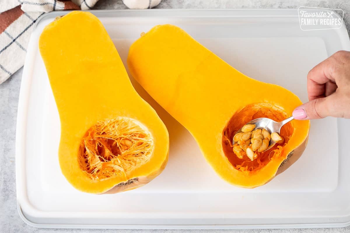Cutting board with a butternut squash cut in half with a spoon scooping out seeds from center.