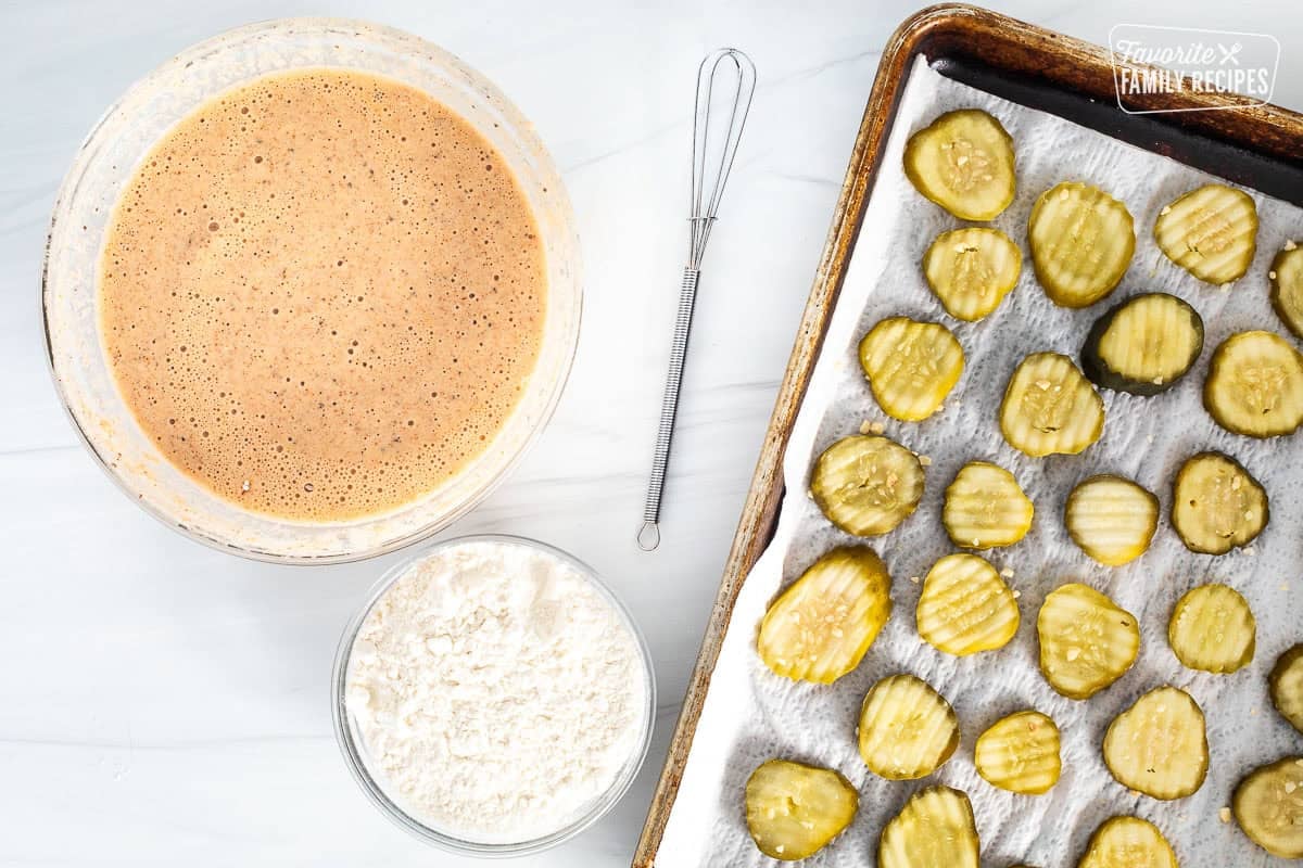 Sheet pan with pickle chips next to flour bowl and batter bowl with whisk.