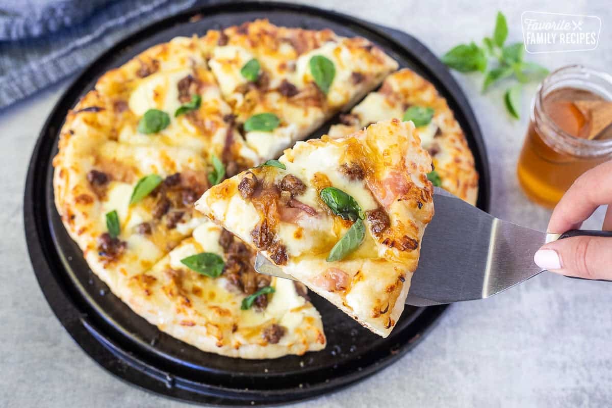 Slice of Hot honey pizza on a spatula with Alfredo sauce, mozzarella cheese, ricotta cheese, prosciutto, sausage, grilled onions, hot honey and fresh basil.