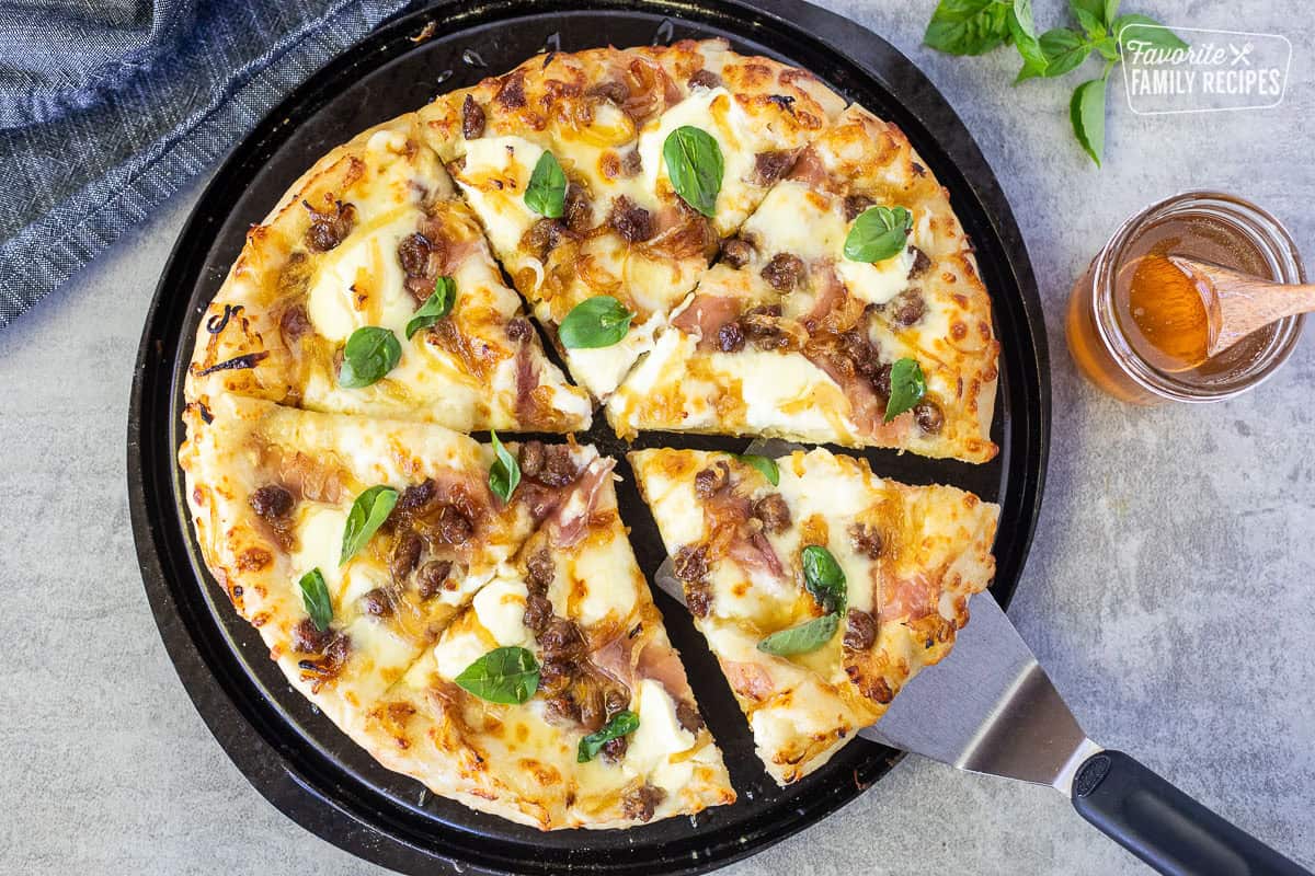 Slice of Hot honey pizza on a spatula with Alfredo sauce, mozzarella cheese, ricotta cheese, prosciutto, sausage, grilled onions, hot honey and fresh basil.