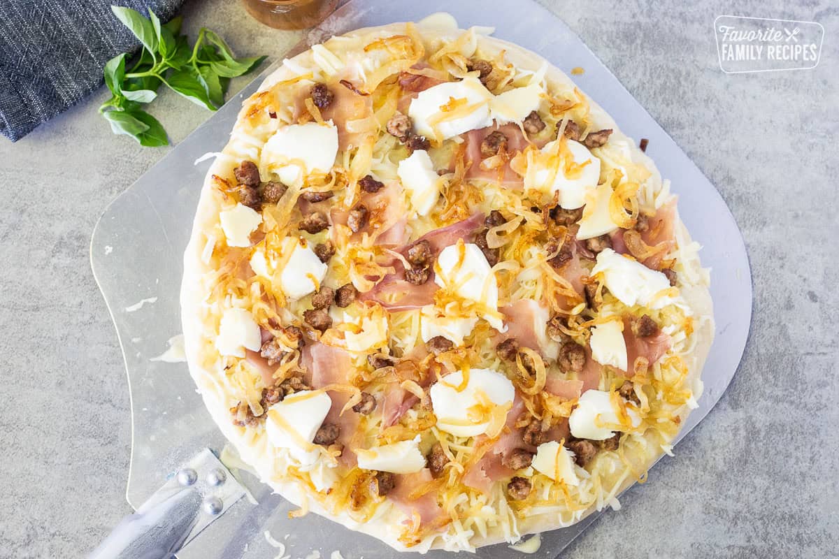 Pizza paddle with baked pizza dough, Alfredo sauce, mozzarella cheese, prosciutto, sausage, ricotta cheese, mozzarella cheese and grilled onions on top.