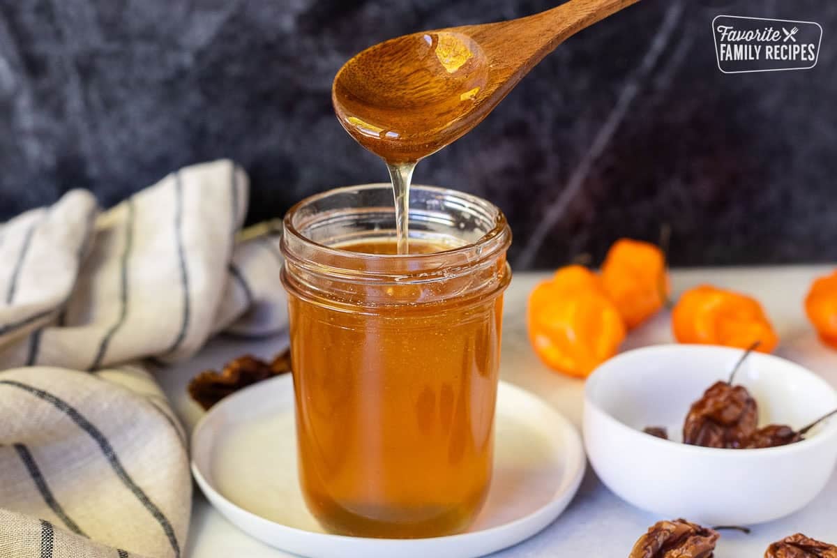 Hot honey in a glass jar with a wooden spoon lifted about and the honey drizzling into the jar. Habanero peppers on the sides.