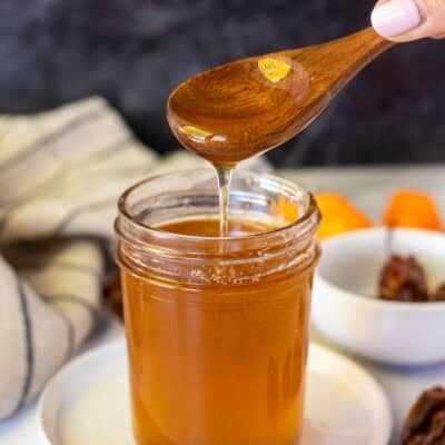 Hot honey in a glass jar with a wooden spoon lifted about and the honey drizzling into the jar. Habanero peppers on the sides.
