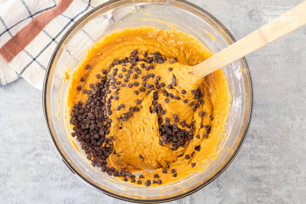 Chocolate chips added to pumpkin bread batter in a large glass mixing bowl with a wooden spoon.