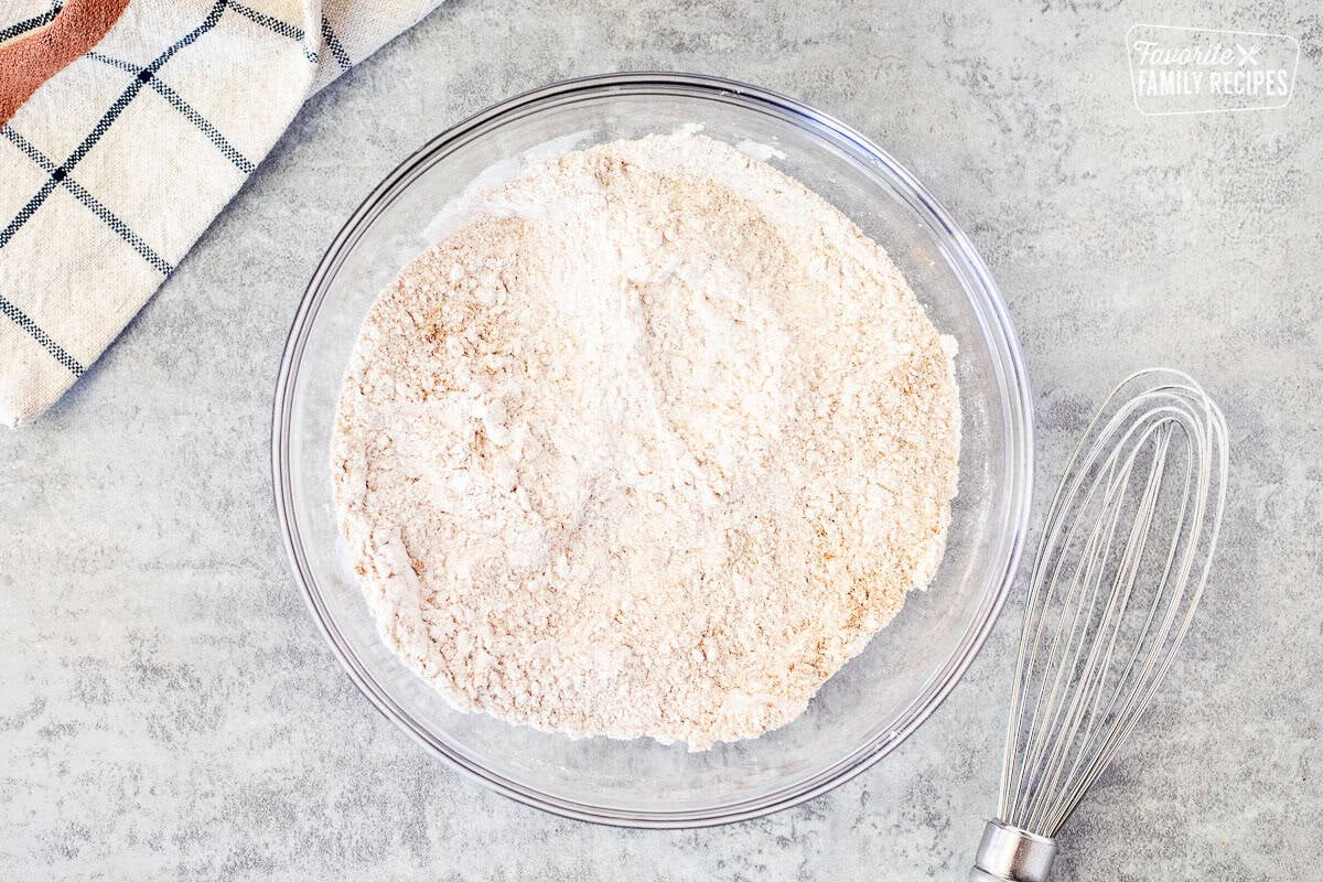 Glass mixing bowl with dry ingredients combined and a whisk on the side.