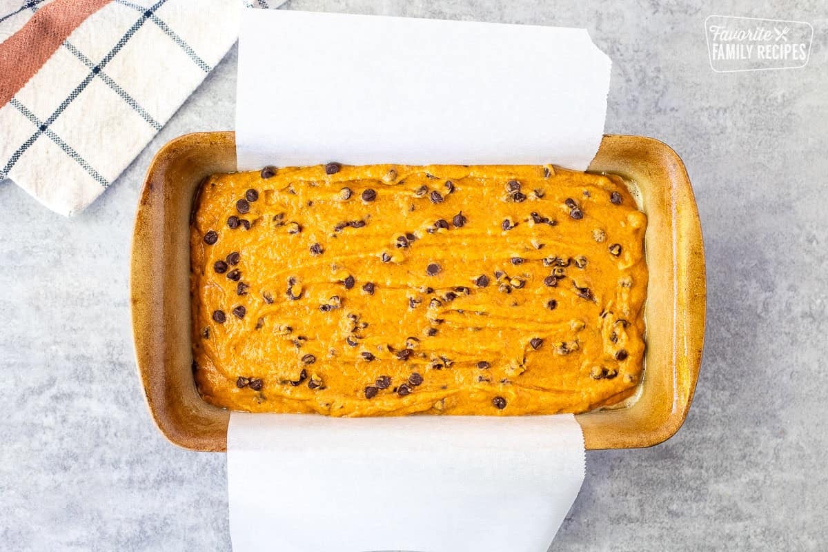 Prepared loaf pan with pumpkin chocolate chip bread batter and parchment paper sticking out the sides.