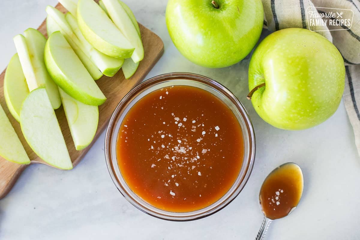 Bowl of salted caramel sauce next to sliced and whole green apples.