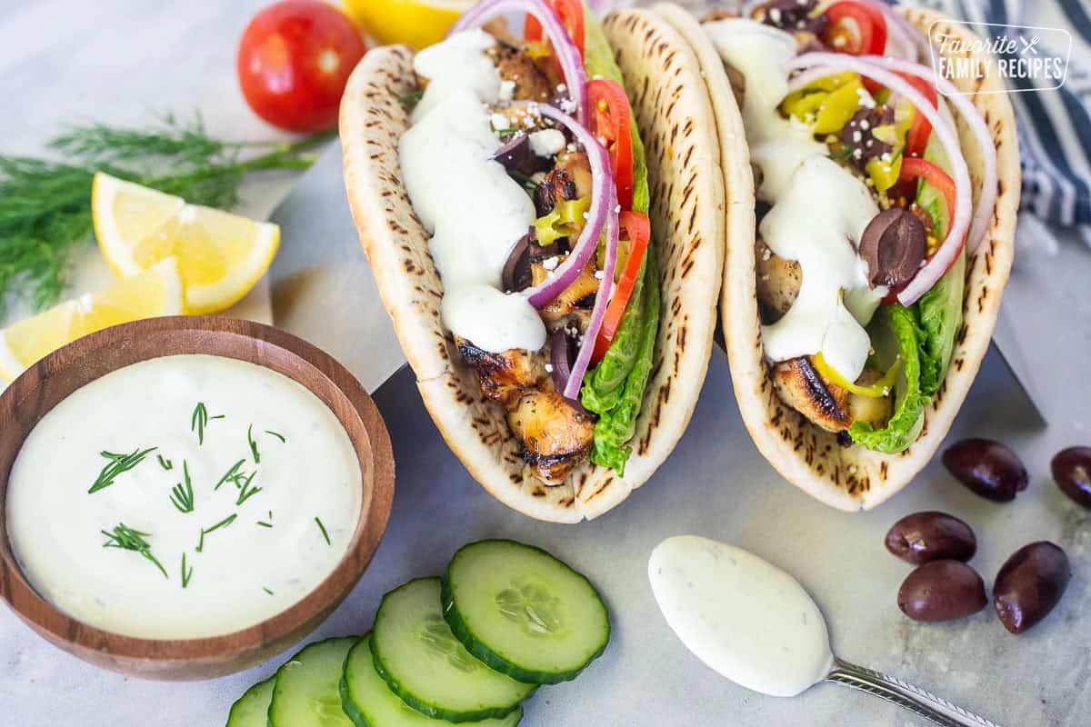 Tzatziki sauce on a chicken gyro with chicken, lettuce, tomatoes, red onion, olives, feta, lemon wedges and peppers. Small bowl and spoon of Tzatziki sauce on the side garnished with fresh dill.