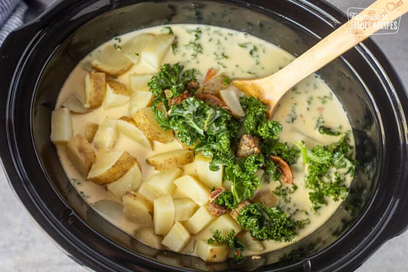 Adding potatoes to a crockpot with kale, sausage, bacon and creamy chicken broth. Wooden spoon resting the crockpot.