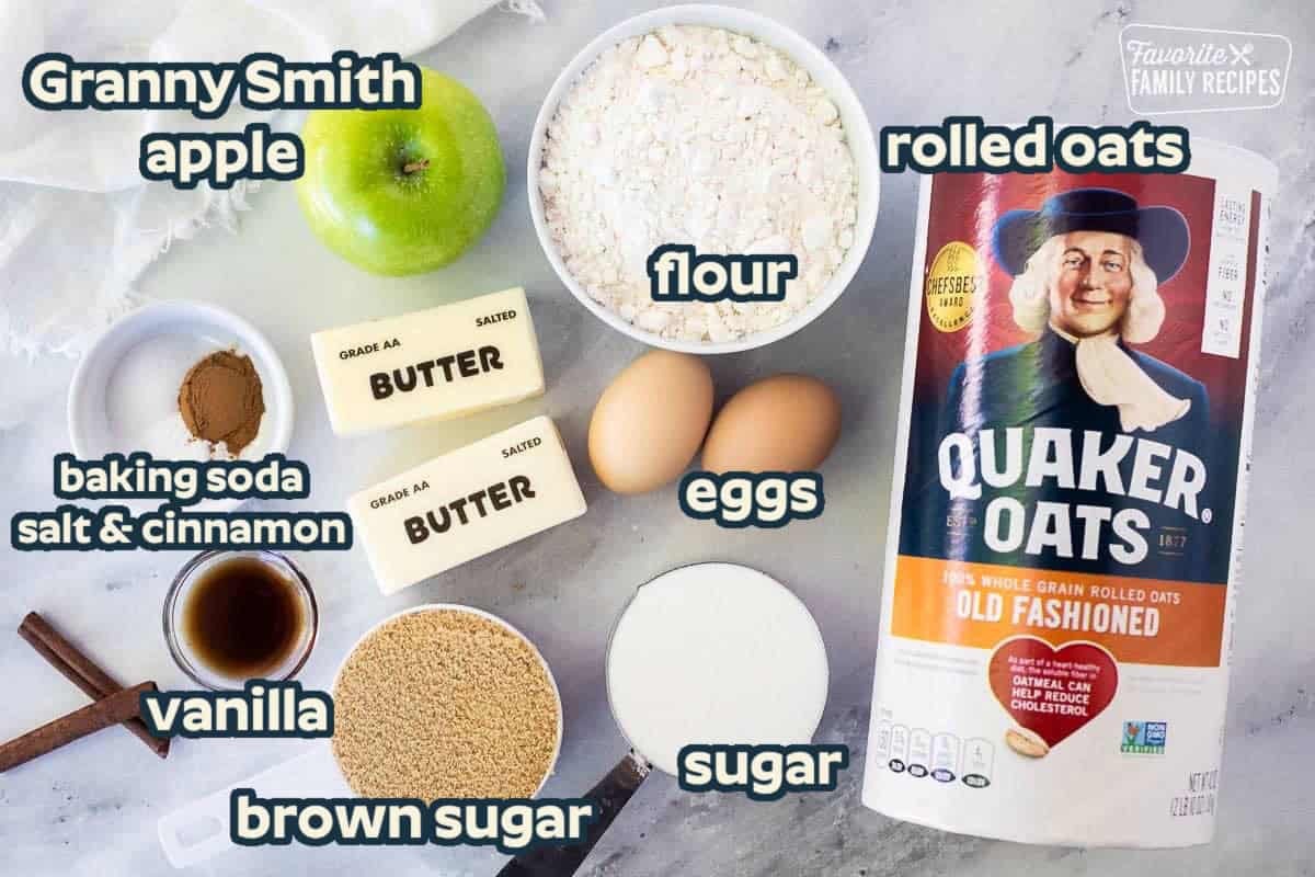 Ingredients to make apple oatmeal cookies including old-fashioned oats, flour, two eggs, sugar, brown sugar, vanilla, cinnamon, salt, baking soda, butter and a green apple.