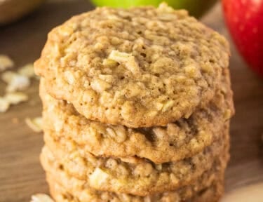 Stack of four Apple oatmeal cookies on a cutting board next to a red and green apple in pieces of oatmeal.