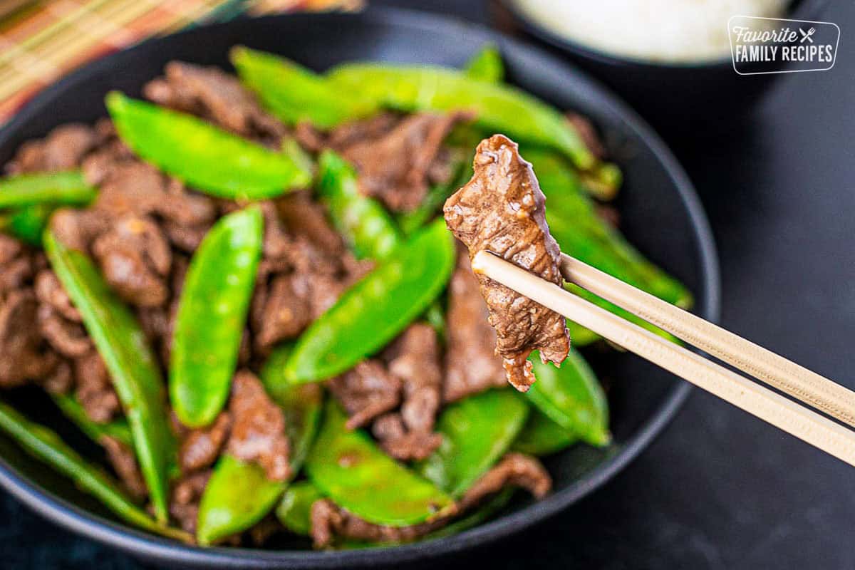 Chopsticks holding up a piece of beef from the bowl of beef and snow peas.