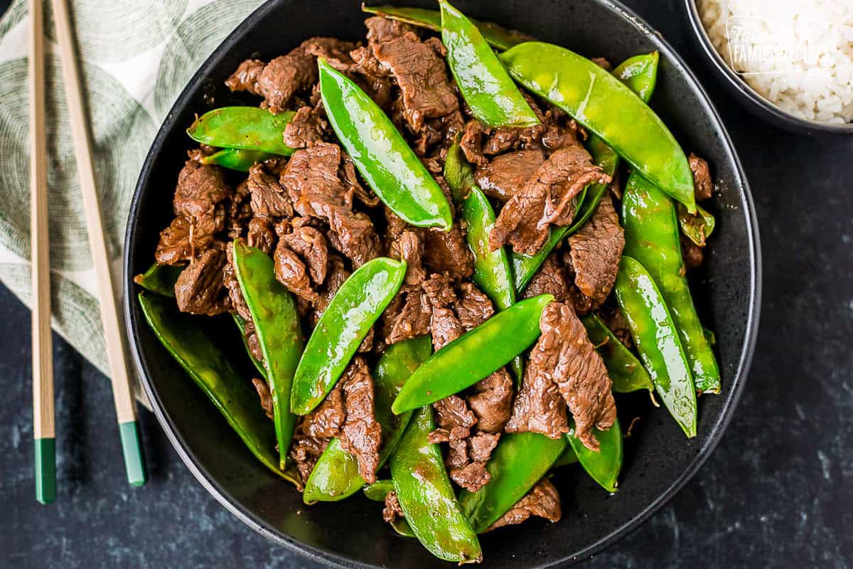 Beef and snow peas in a bowl next to a bowl of rice and chopsticks.