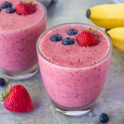 Two glasses of berry banana smoothie garnished with fresh strawberries and blueberries on top.