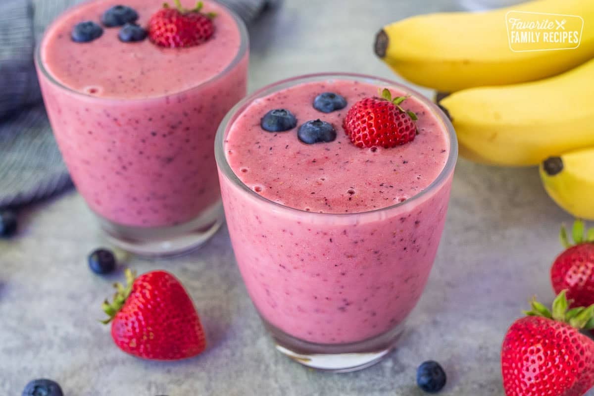 Two glasses of berry banana smoothie garnished with fresh strawberries and blueberries on top.