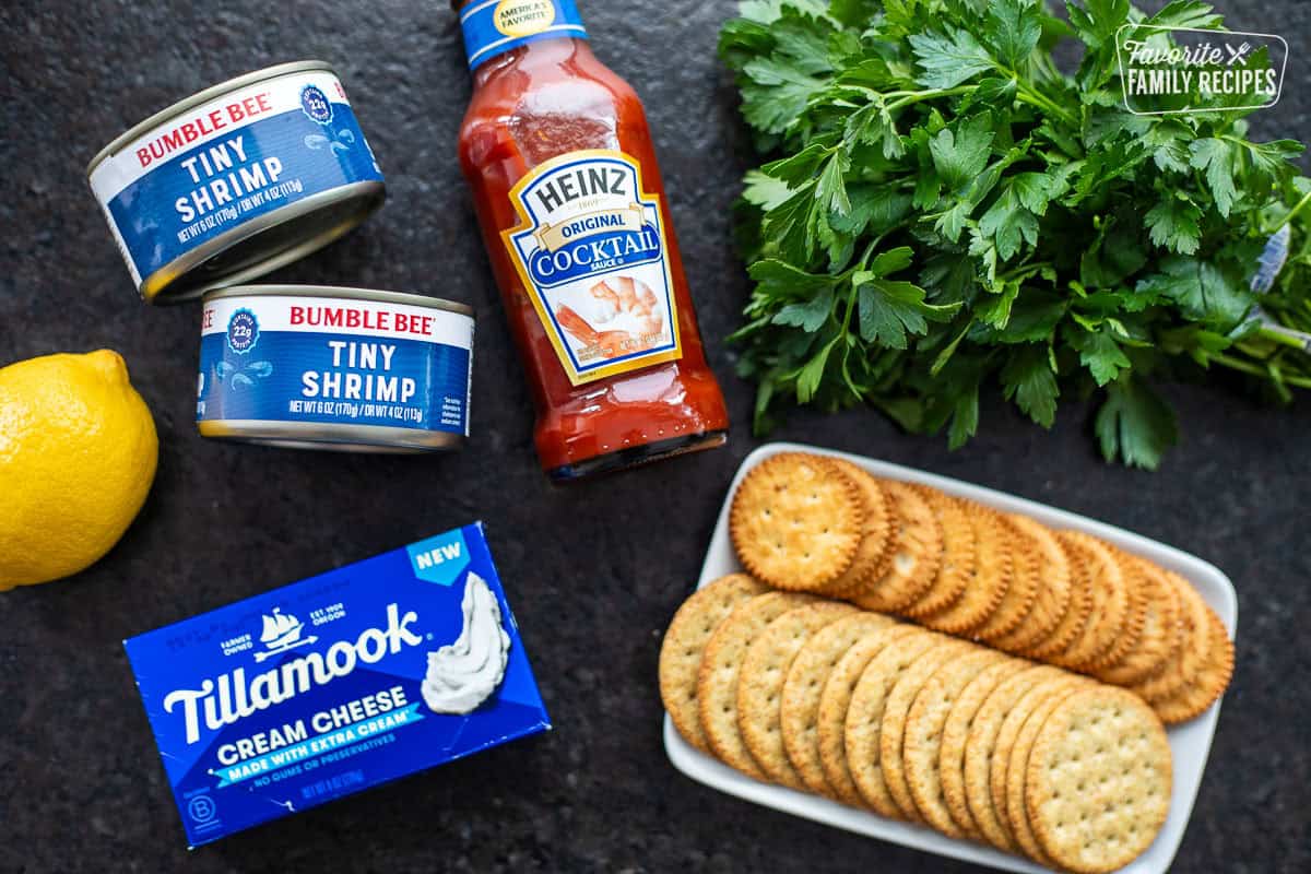 Ingredients to make cream cheese shrimp dip including cream cheese, cans of tiny shrimp, lemon, cocktail sauce, parsley and crackers.