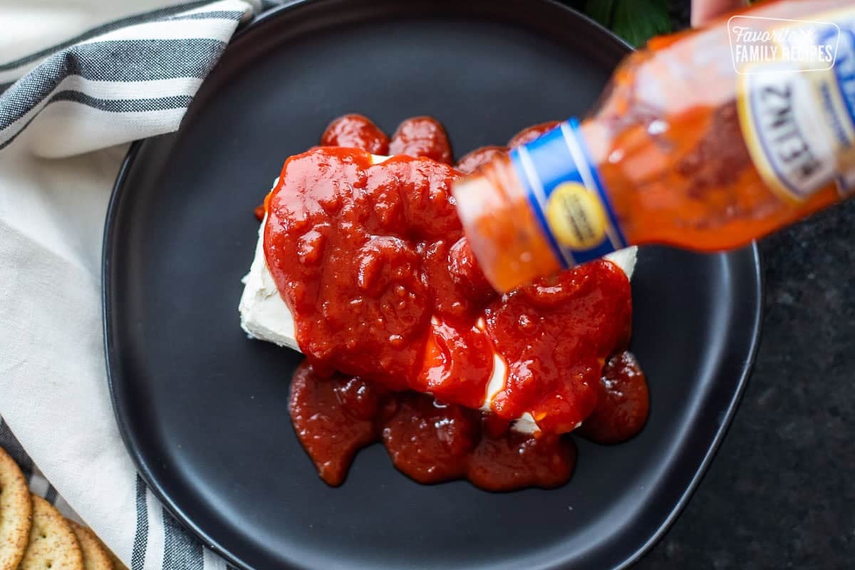 Pouring cocktail sauce on top a brick of cream cheese on a plate.