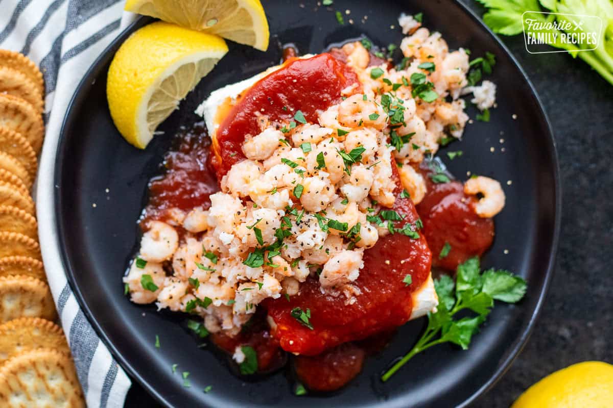 Plate with cream cheese shrimp dip consisting of cream cheese, cocktail sauce, tiny shrimp and garnished with parsley. Crackers on the side.