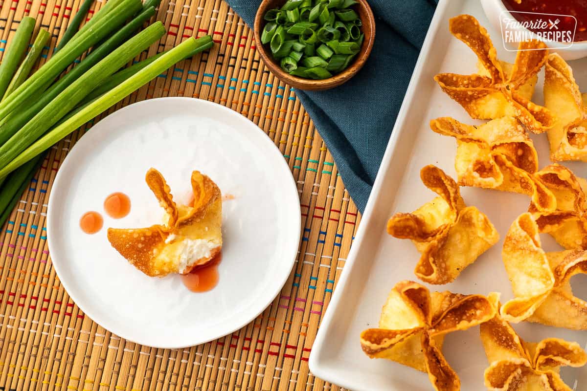Cream cheese wontons on a platter next to a bowl of sweet-and-sour sauce. Small plate with a cut opens cream cheese wonton dipped in sweet-and-sour sauce.