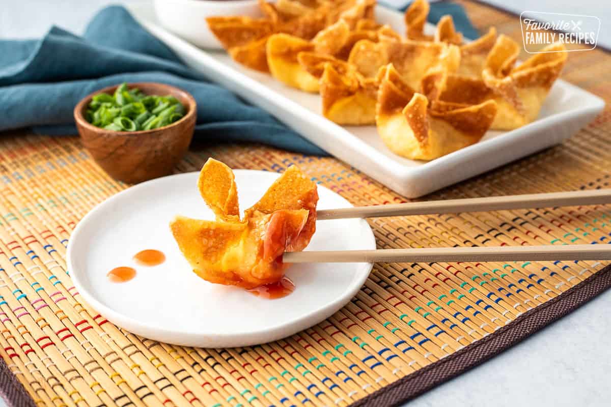 Chopsticks holding a cream cheese wonton dipped in sweet-and-sour sauce on a small plate.