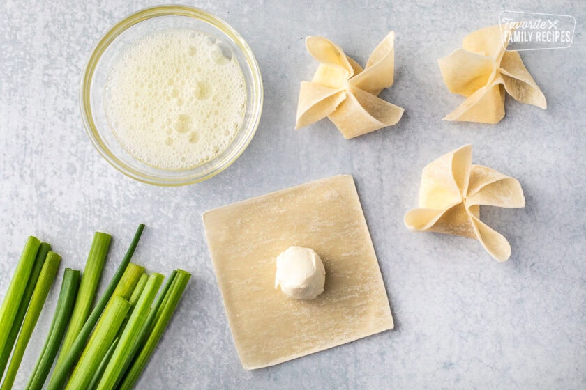 Wrapped cream cheese wonton wrappers next to a laid out wonton wrapper and a bowl of cream cheese in the center. Small bowl of egg wash on the side.