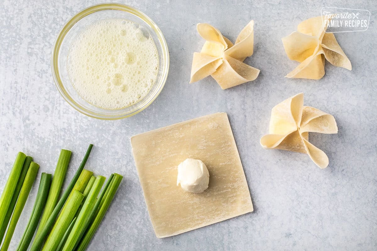 Wrapped cream cheese wonton wrappers next to a laid out wonton wrapper and a bowl of cream cheese in the center. Small bowl of egg wash on the side.