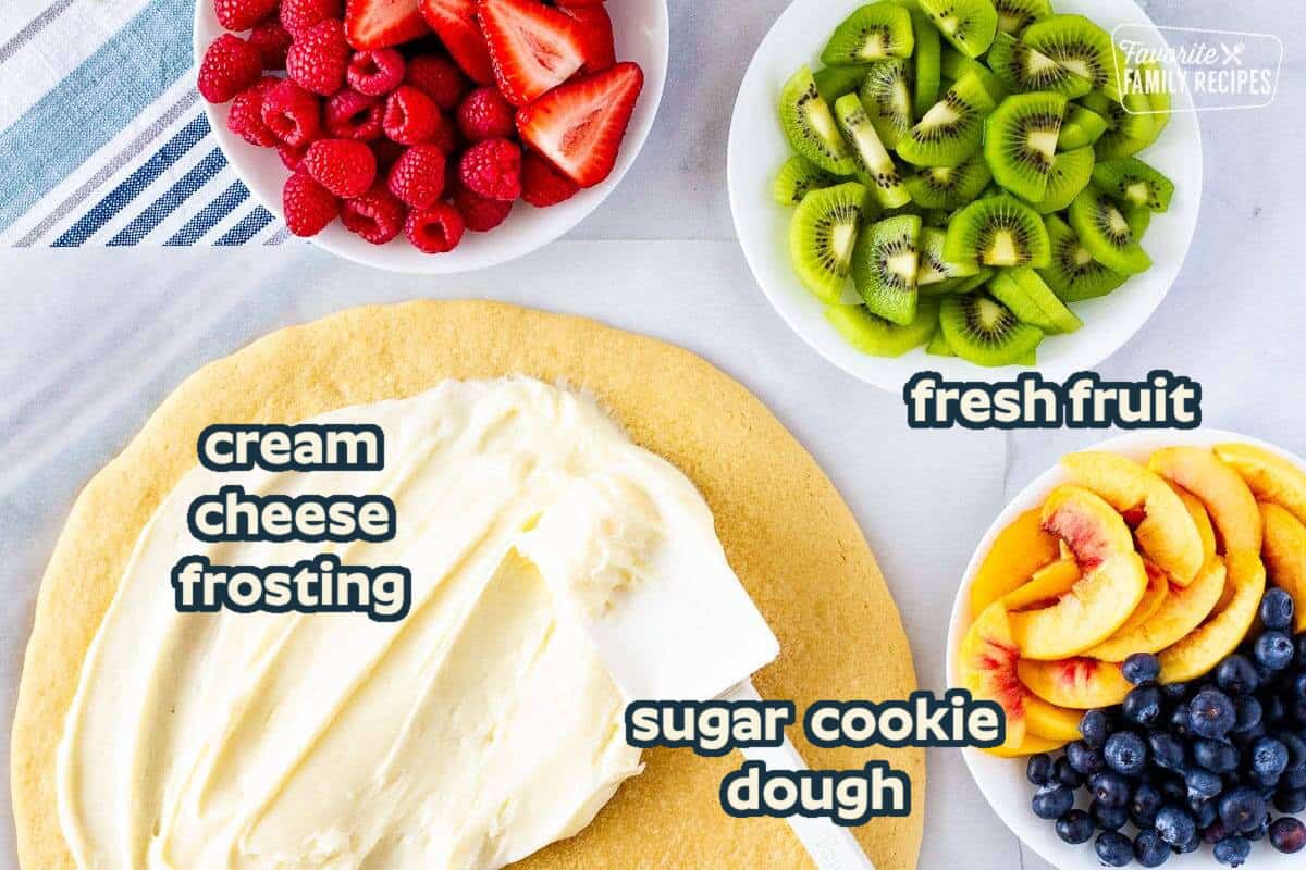 Cookie pizza with a spatula spreading cream cheese frosting and three bowls of fresh fruit on the side.