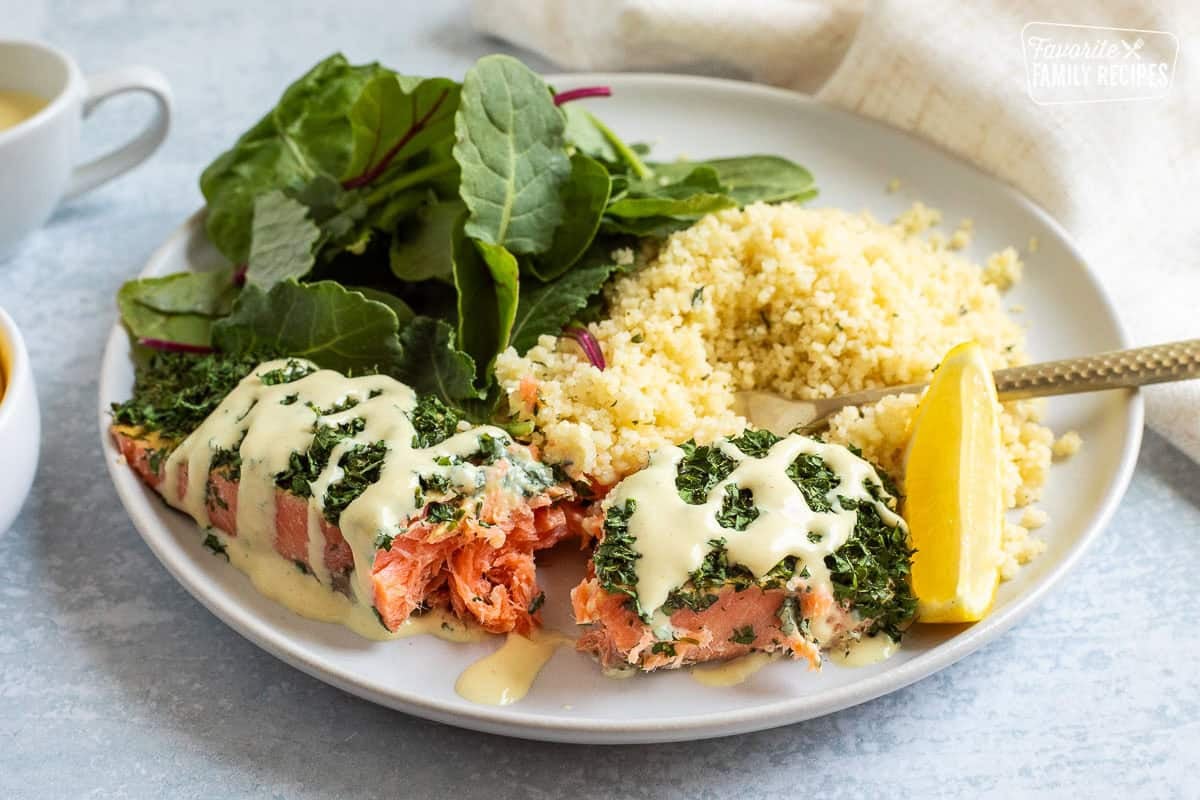 Herbed salmon with dressing cut up with a fork on a plate with salad and couscous.