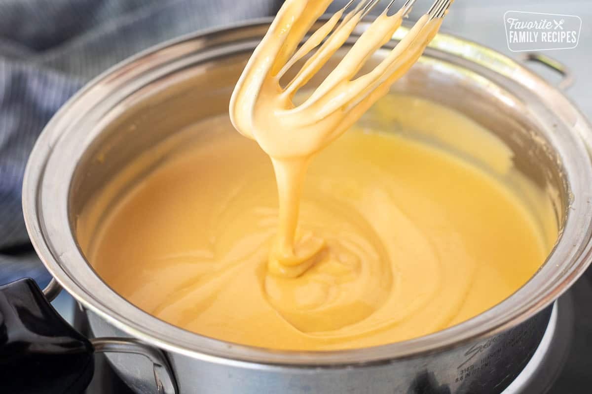 Drizzling cheddar cheese sauce from a whisk into a saucepan.